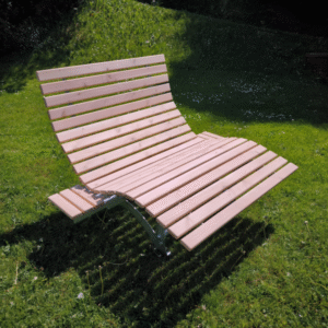 Eine moderne Holzliege, auch Waldsofa oder Wellenliege genannt, mit ausklappbaren Tischen, steht auf einer grünen Wiese in der Sonne. Die Liege ist aus hellen Holzlatten gefertigt und hat ein geschwungenes Design, das zum Entspannen einlädt. Das Untergestell ist aus Stahl.